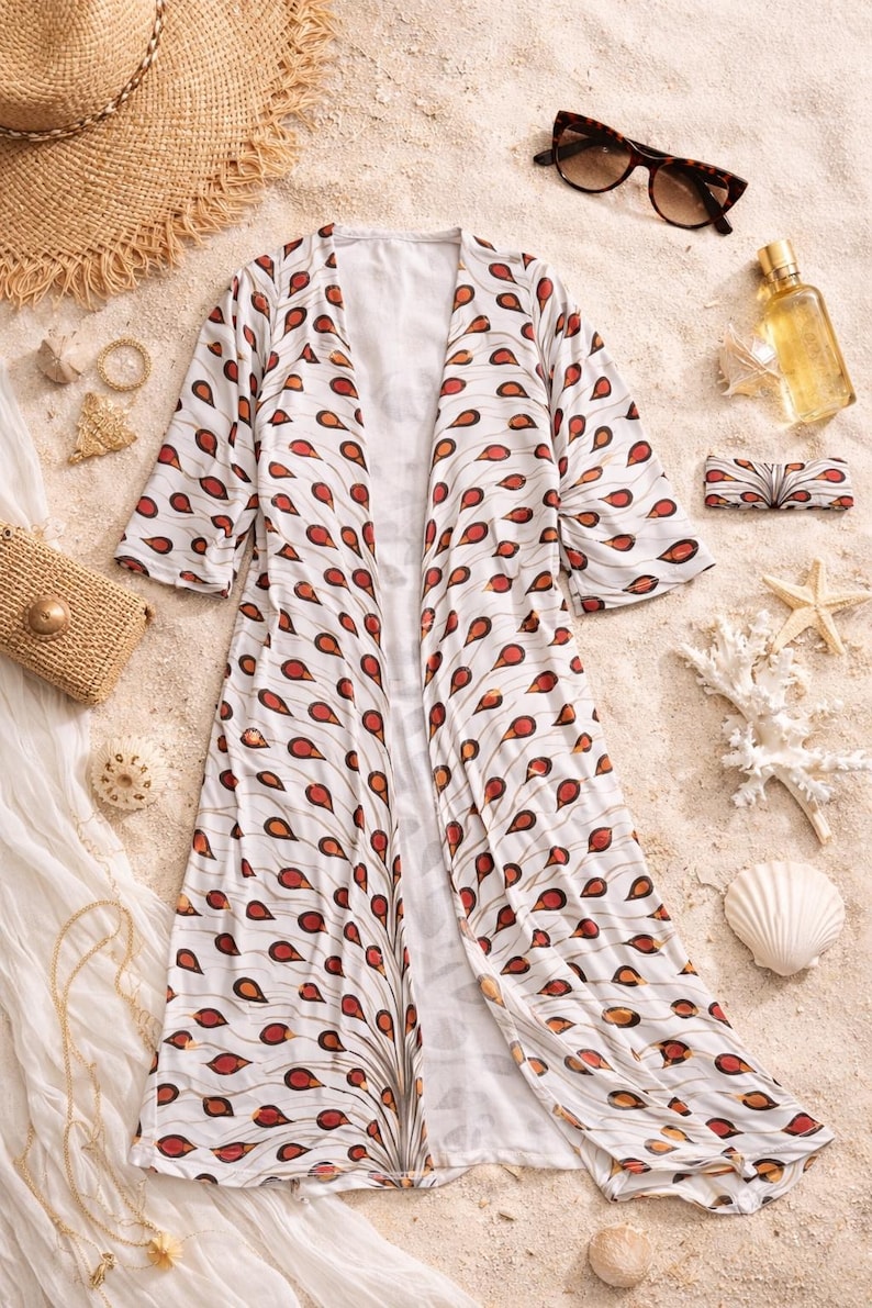 May include: A white kimono with a red and orange teardrop design, displayed on sand with a straw hat, sunglasses, and accessories. The lightweight, flowing garment is ideal for beachwear or a summer cover-up. The kimono's length is approximately 130 cm.