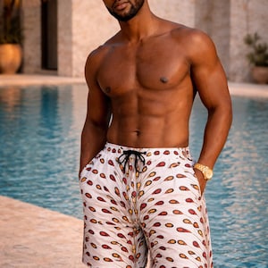 May include: Swim shorts with a white background and a repeating pattern of red, orange, and yellow teardrop shapes. The shorts have a black drawstring. A person is wearing sunglasses and a gold watch.