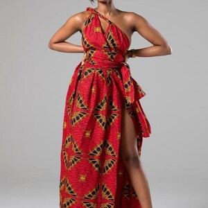 May include: A woman wearing a red and gold patterned maxi dress with a one-shoulder neckline and a thigh-high slit. The dress has a fitted bodice and a flowing skirt. She is wearing gold heels.