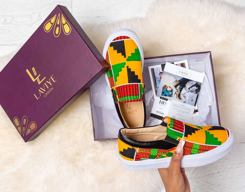 May include: A pair of slip-on sneakers with a colorful geometric pattern in yellow, green, red, and black. The shoes have white soles and black leather trim. They are displayed in an open purple box with gold accents and the brand name "LAVIYE LONDON".