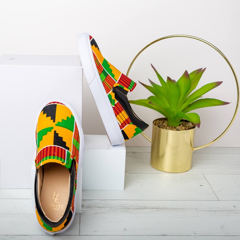 May include: Slip-on sneakers with a vibrant African print in yellow, green, red, and black. The shoes have white soles and black accents. The shoes are displayed on a white surface.