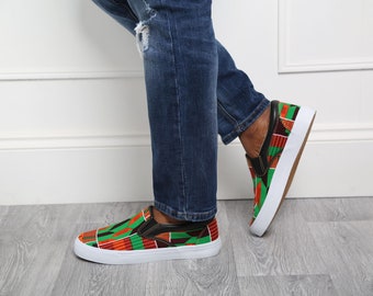 May include: Slip-on sneakers with a vibrant African print in red, green, and black. The shoes have a white sole and black trim. The sneakers are worn with blue denim jeans.