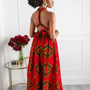 May include: A red and gold maxi dress with a criss-cross back detail. The dress has a floral print and is worn by a model with a matching head wrap.