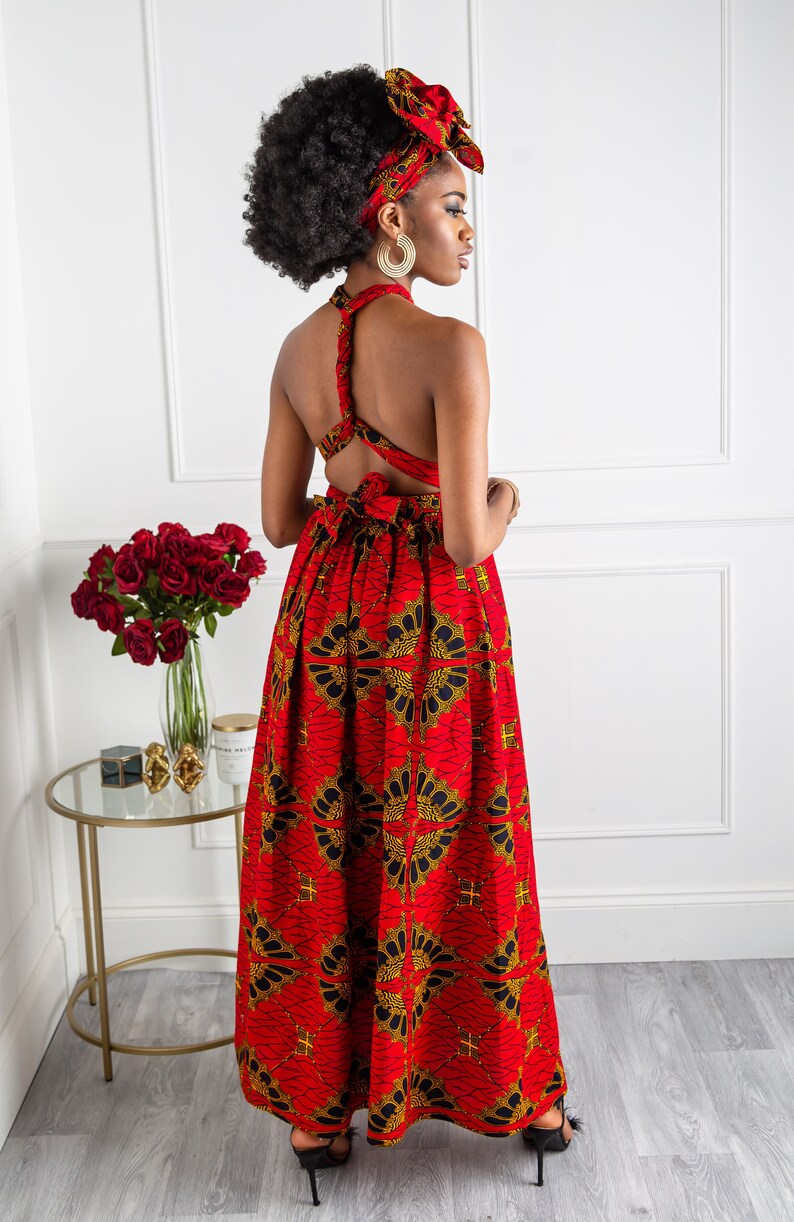 May include: A red and gold maxi dress with a criss-cross back detail. The dress has a floral print and is worn by a model with a matching head wrap.