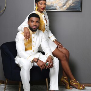 May include: A white long-sleeved shirt with a gold textured panel on the chest and gold buttons, paired with matching white trousers. The shirt features a high collar and gold trim on the cuffs. The outfit is worn with brown patterned loafers.