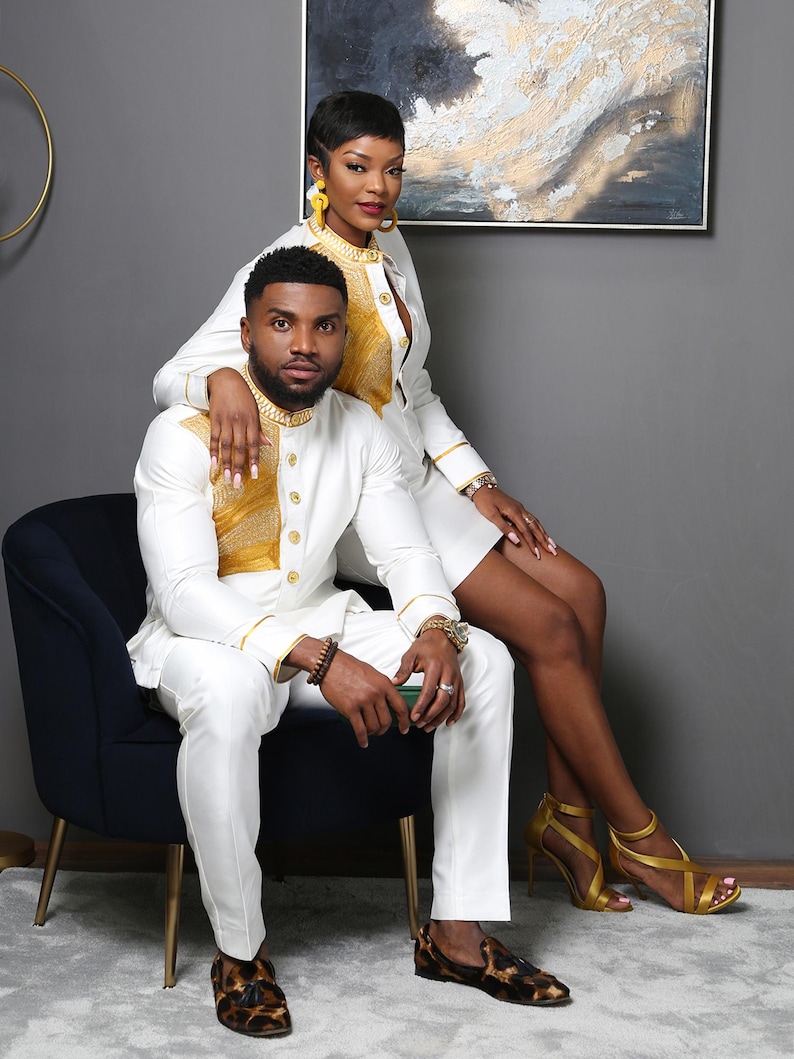 May include: A white long-sleeved shirt with a gold textured panel on the chest and gold buttons, paired with matching white trousers. The shirt features a high collar and gold trim on the cuffs. The outfit is worn with brown patterned loafers.