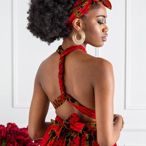 May include: A woman wearing a red and black patterned dress with a halter neckline and a matching head wrap. The dress has a low back and is tied at the waist. She is wearing large gold hoop earrings.