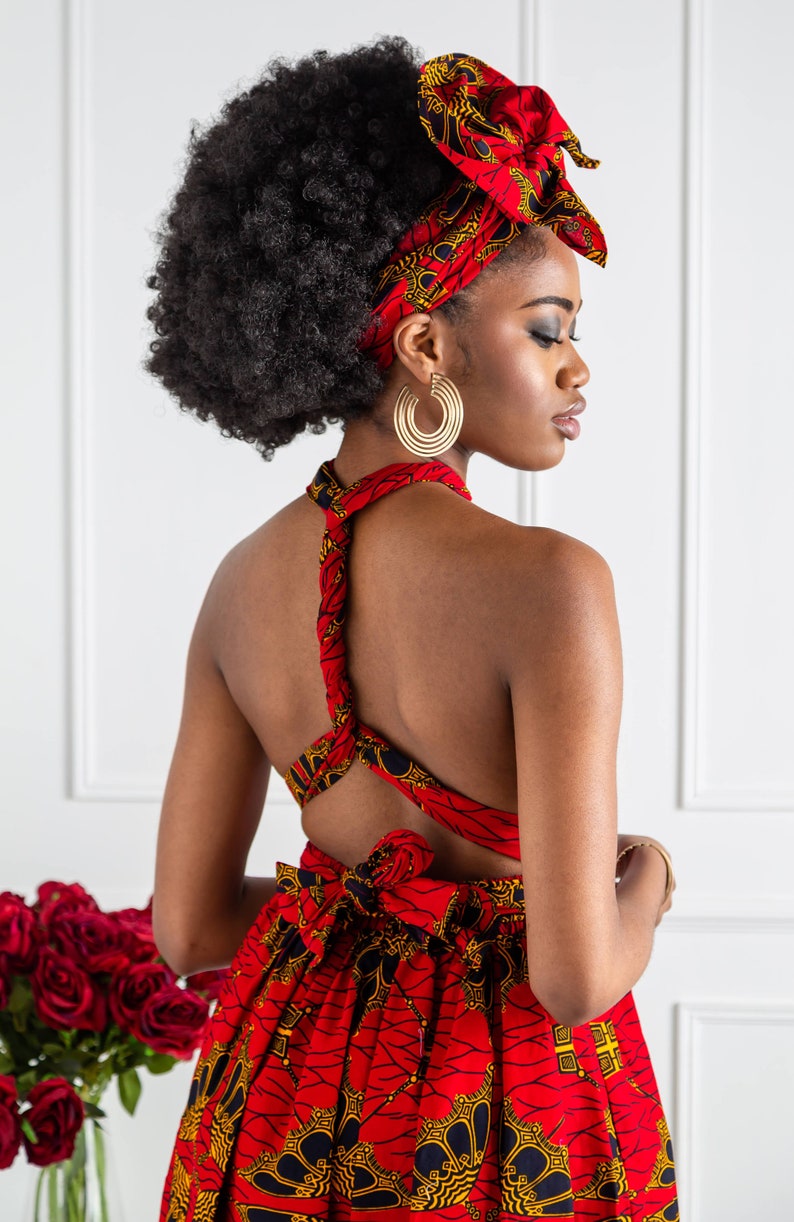 May include: A woman wearing a red and black patterned dress with a halter neckline and a matching head wrap. The dress has a low back and is tied at the waist. She is wearing large gold hoop earrings.