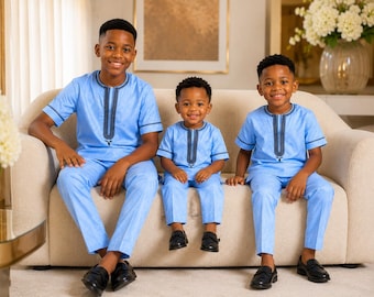 May include: Three children wearing matching light blue short-sleeved shirts and trousers. The shirts feature a round neckline with a vertical dark blue and white embroidered placket down the centre and matching trim on the sleeve cuffs.