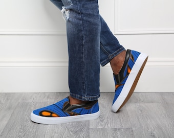 May include: Blue slip-on sneakers with a vibrant orange and black pattern. The shoes have a white sole and a black leather accent on the heel. The sneakers are paired with blue denim jeans.