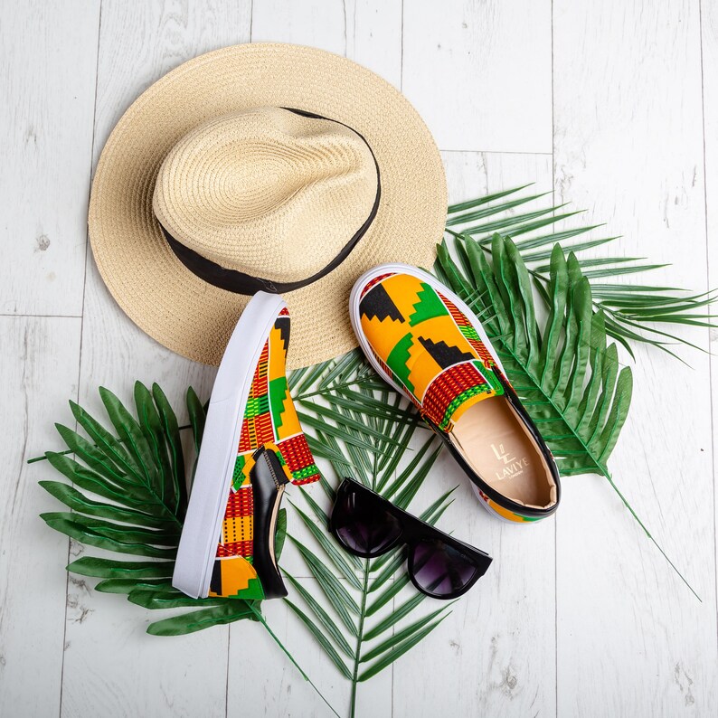 May include: A pair of slip-on shoes with a colorful African print, a straw hat with a black band, and black sunglasses are arranged on a white wooden surface with green palm leaves. The shoes have white soles and black leather accents.