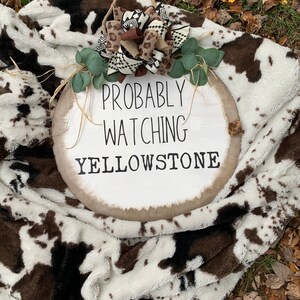 May include: A white round wooden sign with the text "Probably Watching Yellowstone" in black lettering. The sign is decorated with a brown and white patterned bow and green leaves. The sign is sitting on a white and brown cow print blanket.