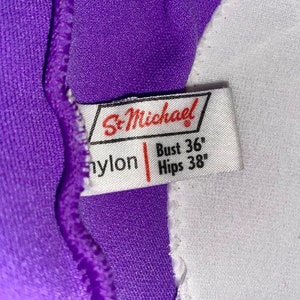 Op de afbeelding: Een wit label met de tekst "St. Michael" in rode letters. Het label bevat ook de tekst "nylon" en de maatgegevens: "Bust 36" en "Hips 38".