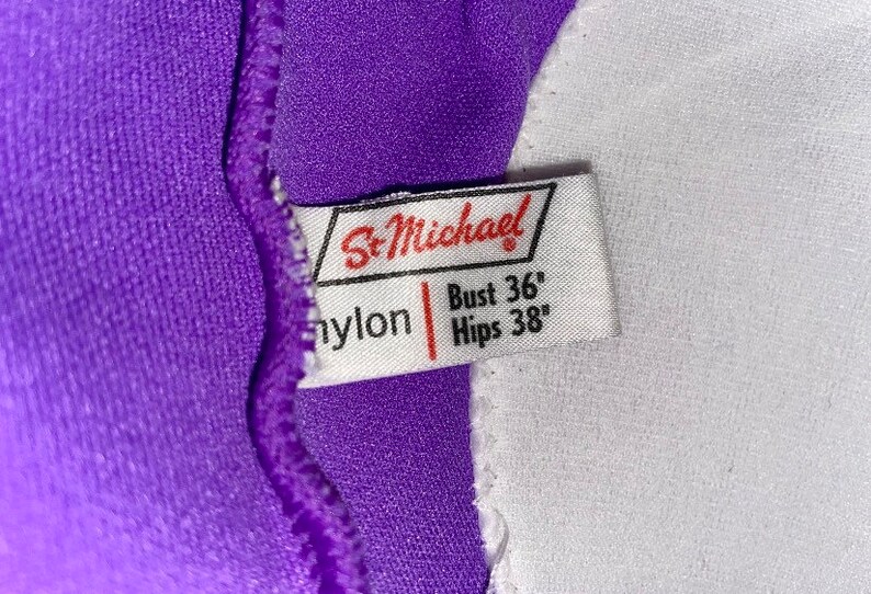 Op de afbeelding: Een wit label met de tekst "St. Michael" in rode letters. Het label bevat ook de tekst "nylon" en de maatgegevens: "Bust 36" en "Hips 38".