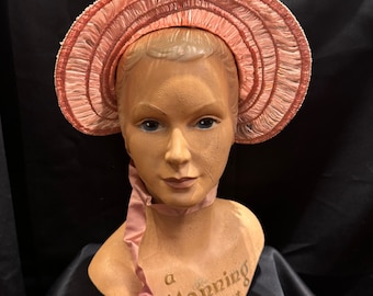 Op de afbeelding: Een roze strohoed met een brede rand en een gerimpelde rand. De hoed zit op het hoofd van een mannequin. De mannequin heeft een roze lint om zijn nek. Op de borst van de mannequin staat de tekst "a Manning hat" gedrukt.