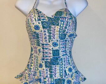 Op de afbeelding: Een vintage halter-badpak met een witte achtergrond en een kleurrijk geometrisch patroon in tinten blauw, groen, geel en paars.