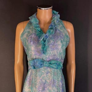 May include: A sleeveless dress featuring a sheer overlay with a floral pattern in shades of teal, blue, and purple. The garment includes a ruffled collar detail around the neckline and a matching fabric bow centered at the waistline.