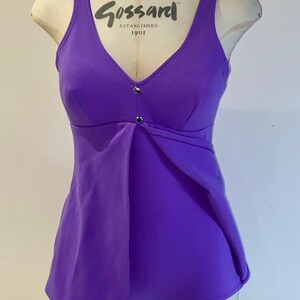 Op de afbeelding: Een paarse badpak met een V-hals en een klein knoopdetail aan de voorkant. Het badpak heeft de tekst "Gossard Established 1901" aan de voorkant.