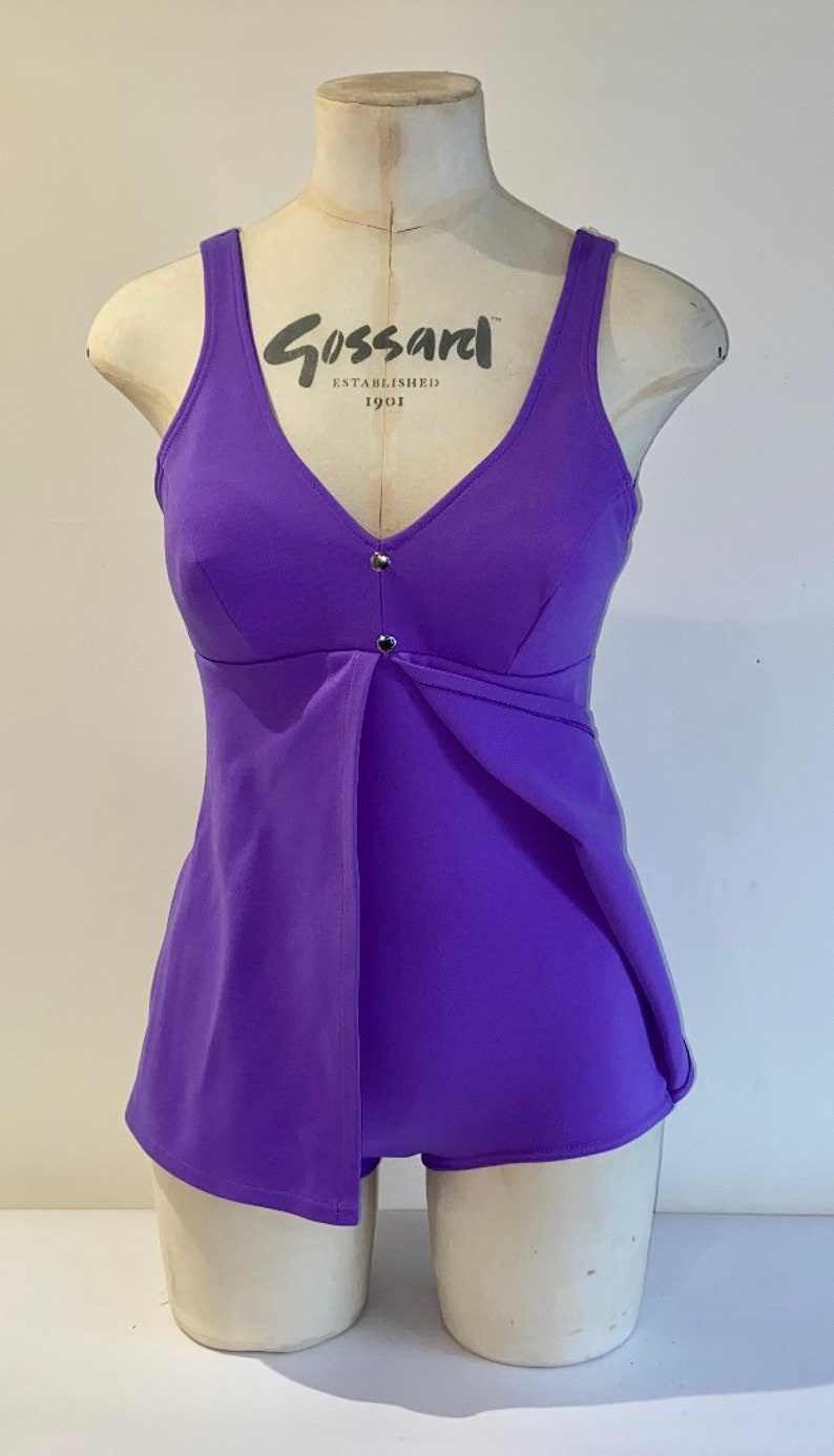 Op de afbeelding: Een paarse badpak met een V-hals en een klein knoopdetail aan de voorkant. Het badpak heeft de tekst "Gossard Established 1901" aan de voorkant.