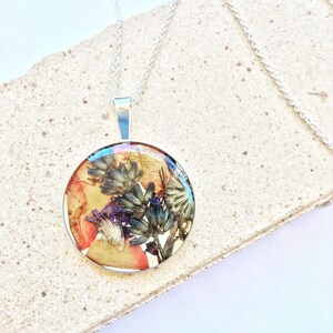 Op de afbeelding: Een zilveren ketting met een ronde hanger met geperste gedroogde bloemen en bladeren ingekapseld in helder hars.
