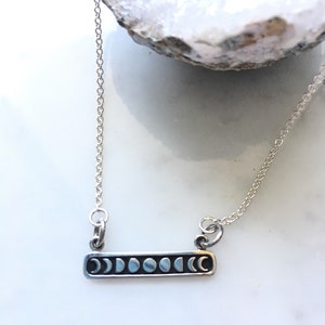 Op de afbeelding: Een zilveren ketting met een staafhanger met acht maanfasen. De ketting ligt op een wit oppervlak.
