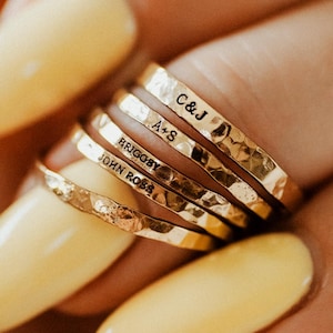 May include: A stack of gold-colored hammered rings, each engraved with a name or initials. The rings are held against a background of yellow-painted fingernails. The inscriptions include "C&J", "A+S", "BRIGGSY", and "JOHN ROSS".