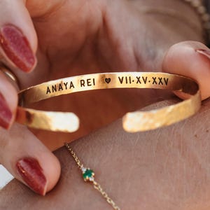 May include: Gold-colored cuff bracelet with engraved text and a heart symbol. The bracelet is held by a person with red nail polish. A delicate gold chain bracelet with a green gemstone is also visible.