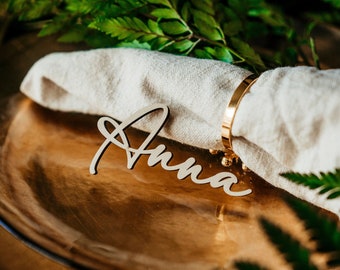 May include: A wooden place card with the name "Anna" written in cursive script. The place card is sitting on a gold plate with a white linen napkin and a gold napkin ring.