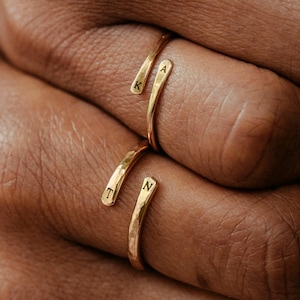 May include: Two gold-toned open rings with a hammered texture. Each ring features two ends, with the letters K and A engraved on the top ring and the letters T and N engraved on the bottom ring. The bands are thin and wrap around the finger.