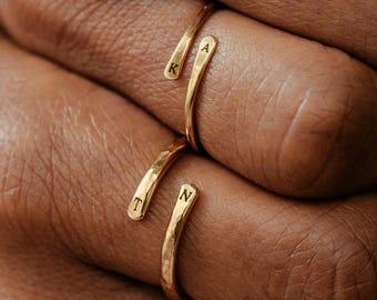 May include: Two gold-toned open rings with a hammered texture. Each ring features two ends, with the letters K and A engraved on the top ring and the letters T and N engraved on the bottom ring. The bands are thin and wrap around the finger.