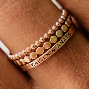 May include: A set of three stacked gold-toned rings. The top ring features a delicate beaded texture, the middle ring has a series of small circular discs, and the bottom flat band is engraved with the text KAIZEN & ANAYA in black lettering.
