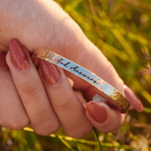 Puede incluir: Un brazalete de metal dorado martillado con la inscripci&oacute;n "God Answers" en una fuente manuscrita. El brazalete se sostiene en una mano con u&ntilde;as rojas pulidas, sobre un fondo borroso de hierba verde.