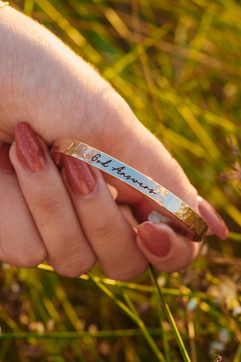 Puede incluir: Un brazalete de metal dorado martillado con la inscripci&oacute;n "God Answers" en una fuente manuscrita. El brazalete se sostiene en una mano con u&ntilde;as rojas pulidas, sobre un fondo borroso de hierba verde.