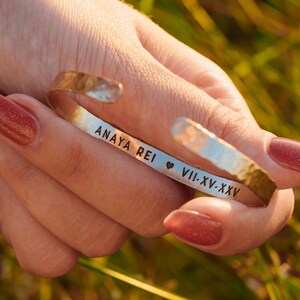 Puede incluir: Una pulsera de pu&ntilde;o plateada con textura martillada. La pulsera tiene grabado el texto "ANAYA REI" seguido de un s&iacute;mbolo de coraz&oacute;n y "VII-XV-XXV". La pulsera se sostiene en una mano con esmalte de u&ntilde;as rojo.