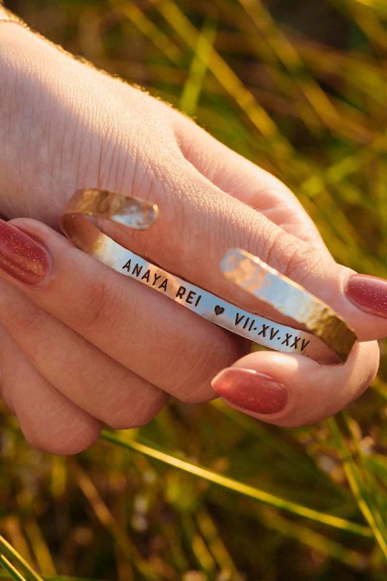 Puede incluir: Una pulsera de pu&ntilde;o plateada con textura martillada. La pulsera tiene grabado el texto "ANAYA REI" seguido de un s&iacute;mbolo de coraz&oacute;n y "VII-XV-XXV". La pulsera se sostiene en una mano con esmalte de u&ntilde;as rojo.