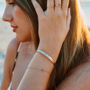 Puede incluir: Una pulsera dorada con una peque&ntilde;a gema verde y una pulsera de pu&ntilde;o plateada con la inscripci&oacute;n "God Answers". Tambi&eacute;n se ve un anillo dorado con una gema verde.