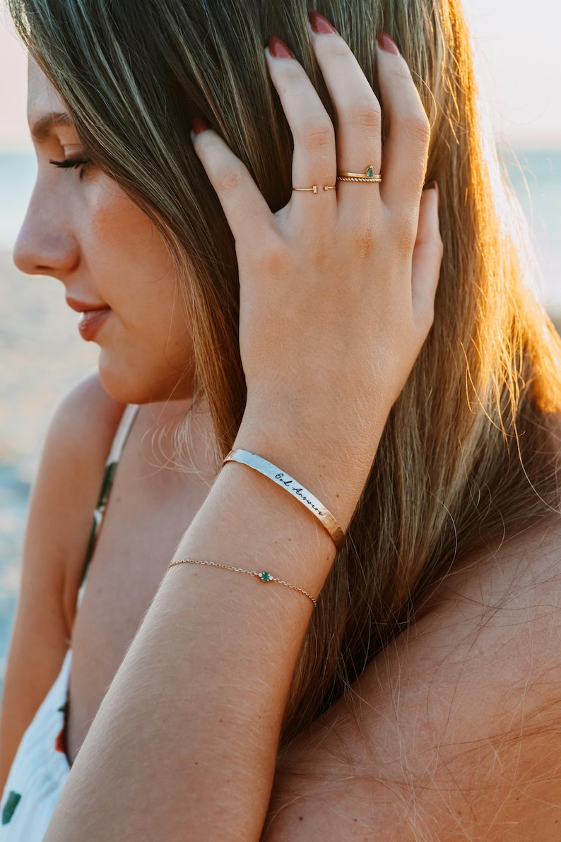 Puede incluir: Una pulsera dorada con una peque&ntilde;a gema verde y una pulsera de pu&ntilde;o plateada con la inscripci&oacute;n "God Answers". Tambi&eacute;n se ve un anillo dorado con una gema verde.