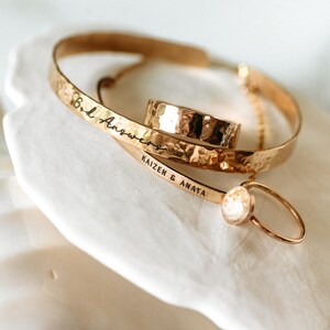 Puede incluir: Una colecci&oacute;n de joyas doradas, que incluye un anillo con acabado martillado, una pulsera con la inscripci&oacute;n "B.L. Answers" y un delicado anillo con una peque&ntilde;a piedra redonda. La pulsera tambi&eacute;n tiene la inscripci&oacute;n "KAIZEN & ANAYA".
