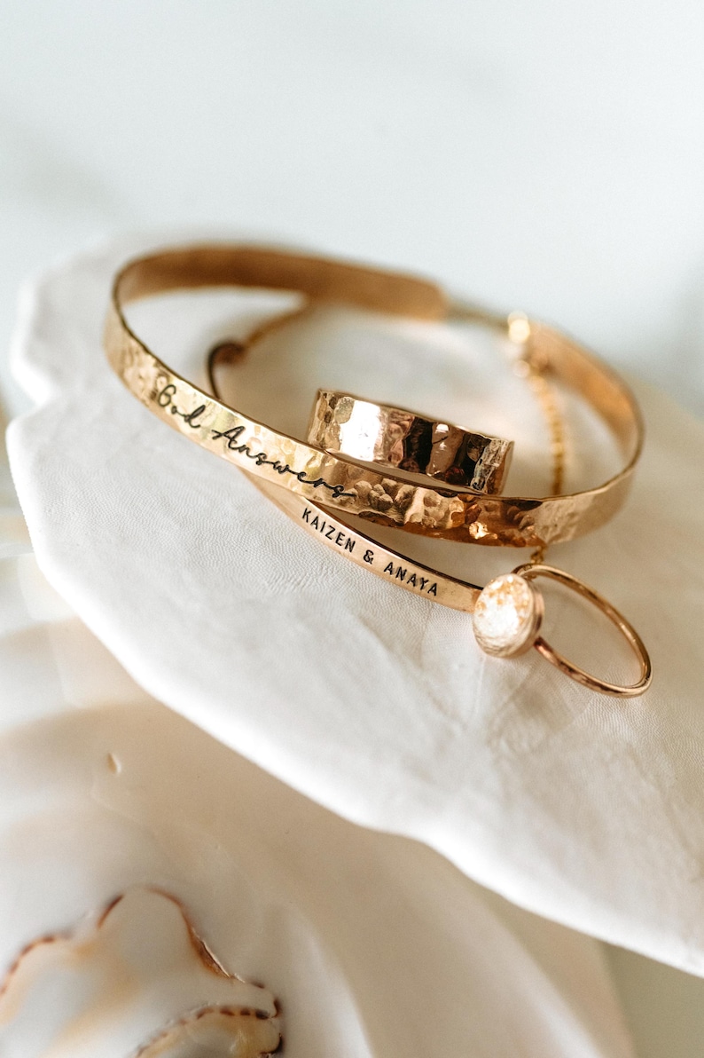 Puede incluir: Una colecci&oacute;n de joyas doradas, que incluye un anillo con acabado martillado, una pulsera con la inscripci&oacute;n "B.L. Answers" y un delicado anillo con una peque&ntilde;a piedra redonda. La pulsera tambi&eacute;n tiene la inscripci&oacute;n "KAIZEN & ANAYA".