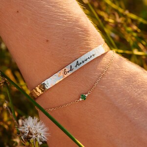 Puede incluir: Una pulsera dorada con la inscripci&oacute;n "God Answers" y una delicada pulsera de cadena dorada con una peque&ntilde;a piedra preciosa verde y diminutas piedras blancas. Las pulseras se llevan en una mu&ntilde;eca, con un fondo borroso de vegetaci&oacute;n.