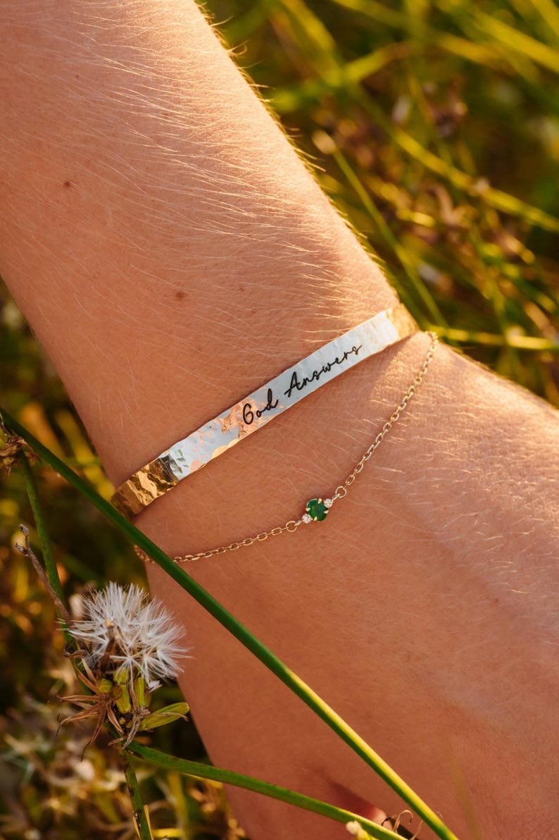 Puede incluir: Una pulsera dorada con la inscripci&oacute;n "God Answers" y una delicada pulsera de cadena dorada con una peque&ntilde;a piedra preciosa verde y diminutas piedras blancas. Las pulseras se llevan en una mu&ntilde;eca, con un fondo borroso de vegetaci&oacute;n.