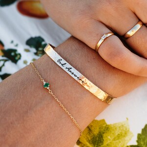 Puede incluir: Una pulsera dorada con la inscripci&oacute;n "God Answers" se usa con una delicada pulsera de cadena dorada con una peque&ntilde;a piedra preciosa verde. Tambi&eacute;n son visibles dos anillos de oro, uno con una peque&ntilde;a piedra rectangular.