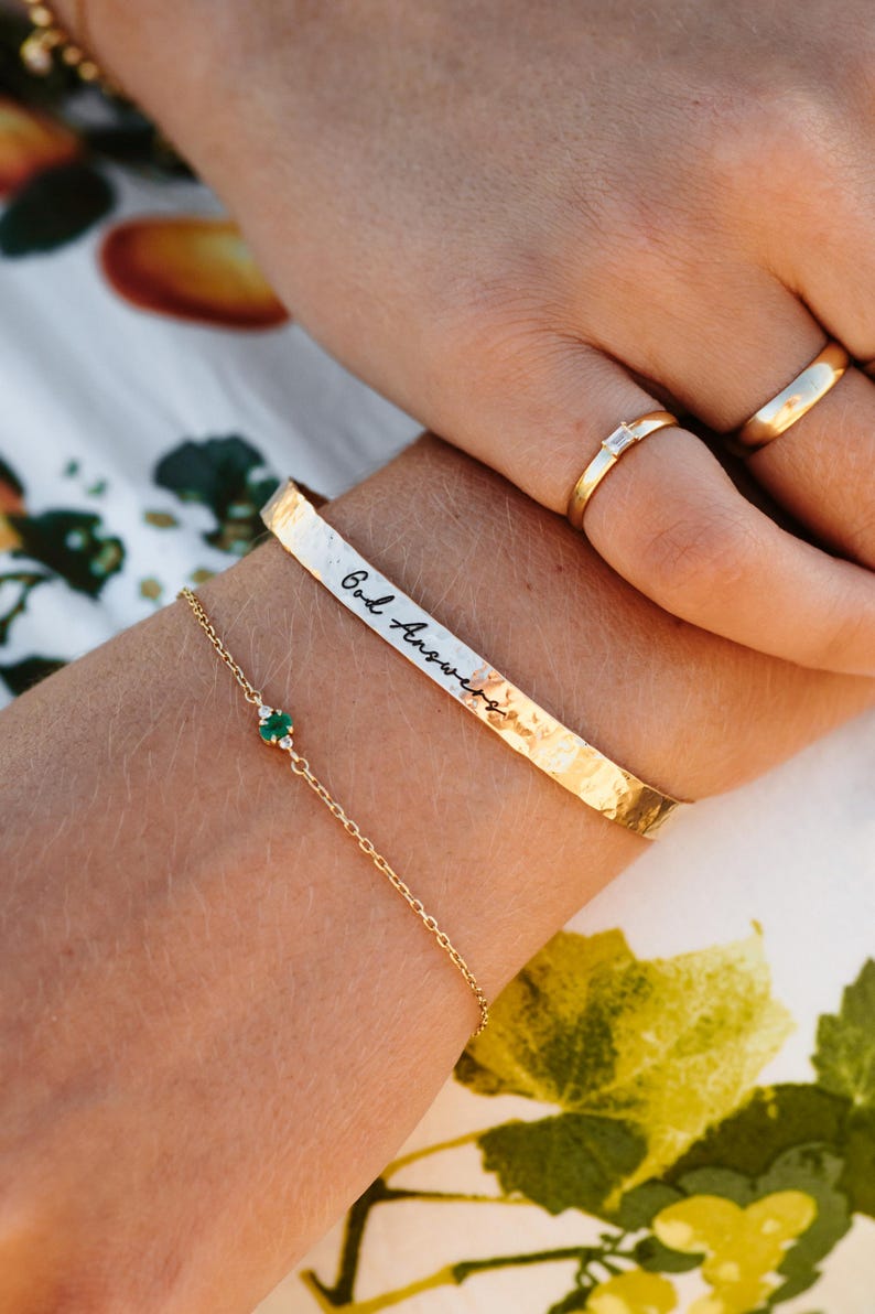 Puede incluir: Una pulsera dorada con la inscripci&oacute;n "God Answers" se usa con una delicada pulsera de cadena dorada con una peque&ntilde;a piedra preciosa verde. Tambi&eacute;n son visibles dos anillos de oro, uno con una peque&ntilde;a piedra rectangular.