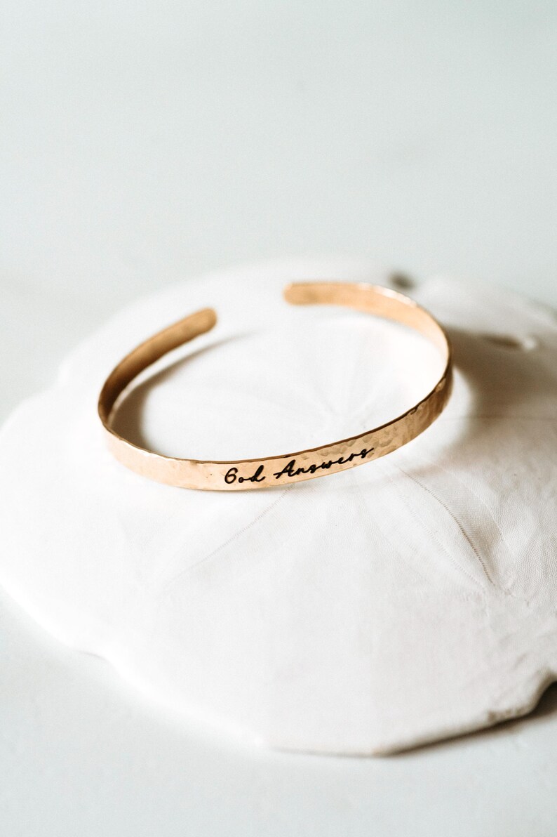 Puede incluir: Un brazalete de color dorado con la inscripci&oacute;n "God Answers" en escritura negra. El brazalete tiene una textura martillada y est&aacute; sobre una superficie blanca texturizada. Joyer&iacute;a.