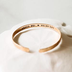 Puede incluir: Una pulsera de pu&ntilde;o dorada con el texto grabado "ANAYA REI" seguido de un s&iacute;mbolo de coraz&oacute;n y n&uacute;meros romanos "VII-XV-XXV". La pulsera abierta se muestra sobre una superficie blanca.
