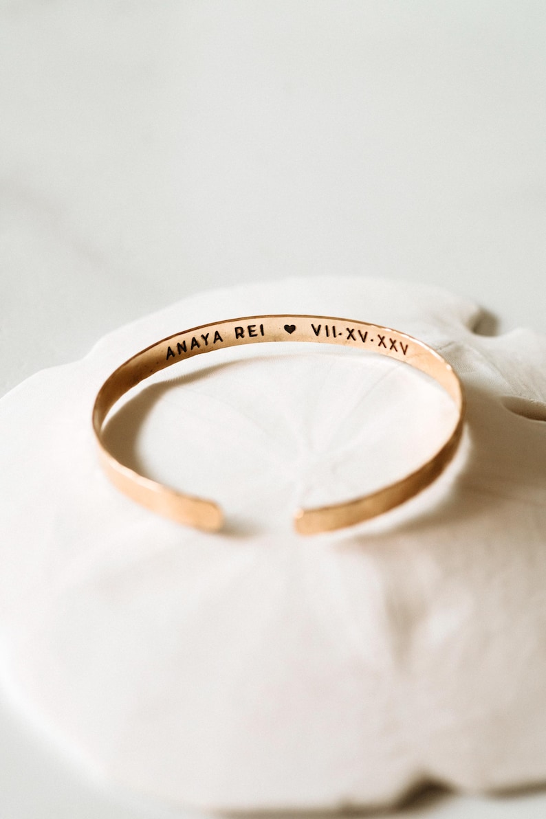 Puede incluir: Una pulsera de pu&ntilde;o dorada con el texto grabado "ANAYA REI" seguido de un s&iacute;mbolo de coraz&oacute;n y n&uacute;meros romanos "VII-XV-XXV". La pulsera abierta se muestra sobre una superficie blanca.