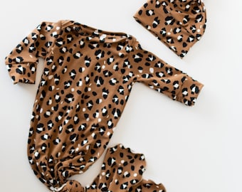 Könnte beinhalten: Ein braunes Baby-Set mit Leopardenmuster in Schwarz und Weiß. Das Set besteht aus einem Strampler mit langen Ärmeln und einem Knoten am unteren Ende. Dazu gehört eine passende Mütze im Beanie-Stil.