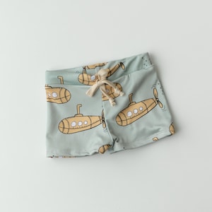 May include: Light blue swim trunks with a repeating pattern of yellow submarines. The trunks have a drawstring closure and are made of a soft, flexible material. The submarines have a propeller and portholes.