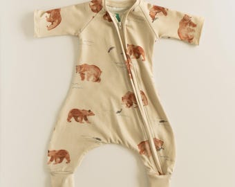 May include: A beige baby sleep sack with a zipper closure and a brown bear print. The sleep sack has long sleeves and a footless design.