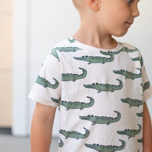 May include: White short-sleeved t-shirt featuring a repeating pattern of green crocodiles. The crocodiles are stylized with curved tails and detailed bodies. The shirt is made of a soft material.